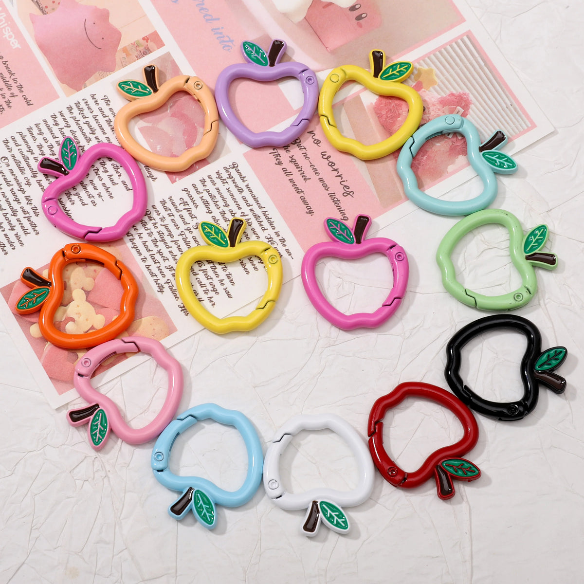 Twelve apple-shaped silicone carabiner clips in various colors (pink, orange, yellow, purple, blue, green, red, black, and white) with green leaf details, arranged on a textured white surface with pink decorative papers underneath.