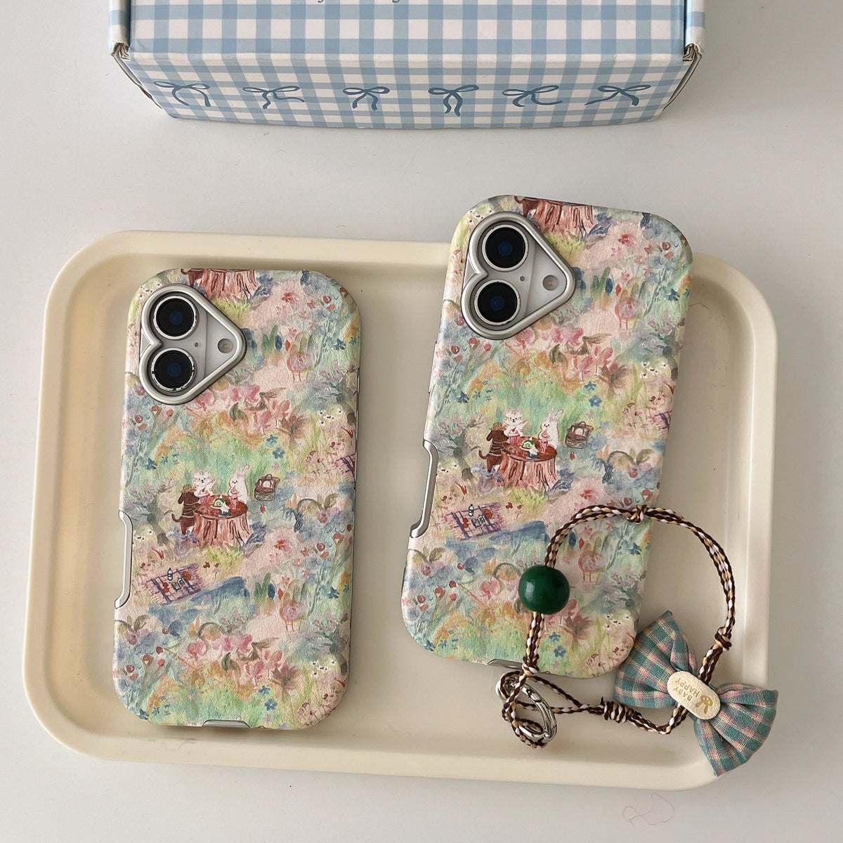 Two floral phone cases displayed on a cream tray, one upright and one lying flat, with a matching blue-and-white gingham box partially visible at the top and a small bow charm attached to the right case.