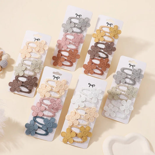 Six carded sets of children's flower hair ties arranged on a light cream background. The sets include different color combinations of cream, pink, brown, yellow, blue, and gray fabric flowers with elastic bands.