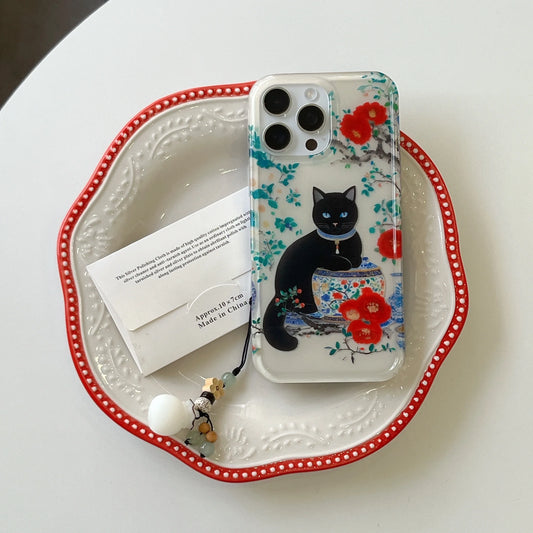 A clear phone case featuring a black cat sitting by a fishbowl with red flowers, displayed on a white ceramic plate with a red rim. A white envelope card and a small charm are placed next to the case.