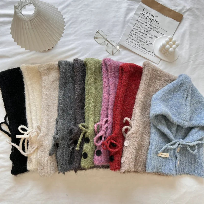 A set of fuzzy knit hooded cardigans or sweaters laid flat on a white fabric surface, shown in multiple colors including black, cream, gray, sage green, pink, red, beige, and light blue. Each piece has a drawstring hood and front buttons.