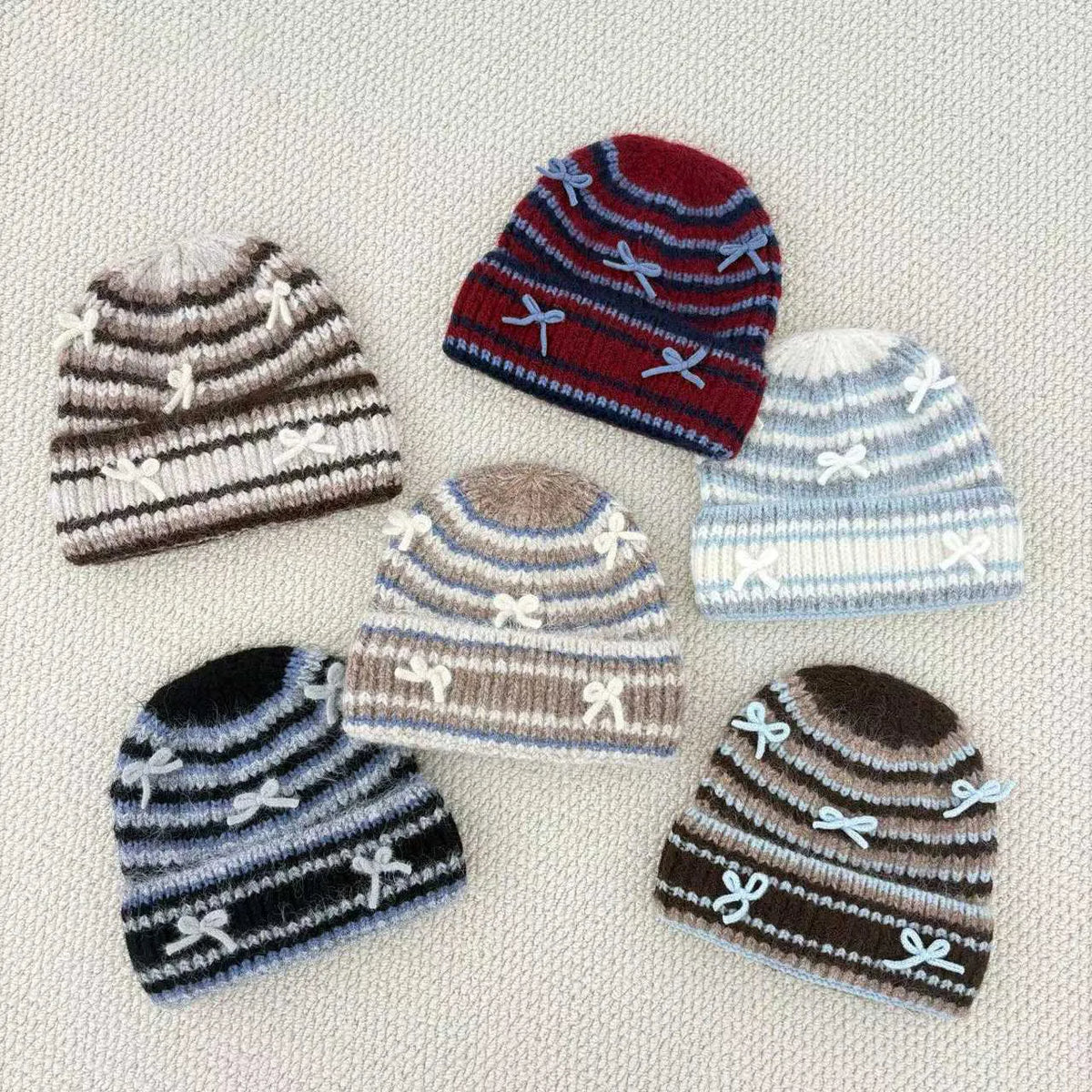 Five striped knit beanie hats arranged on a light beige textured fabric background. The hats feature horizontal stripes in various color combinations including brown, red, blue, gray, and black, with white embroidered bow details and a white pom-pom on two of them.