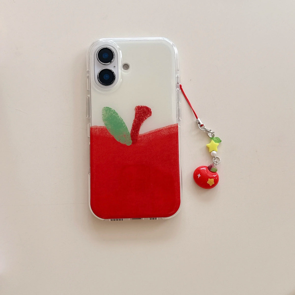 A clear phone case featuring a painted red apple design with green and red leaf details, shown on a phone next to a red and yellow charm strap on a light beige background.