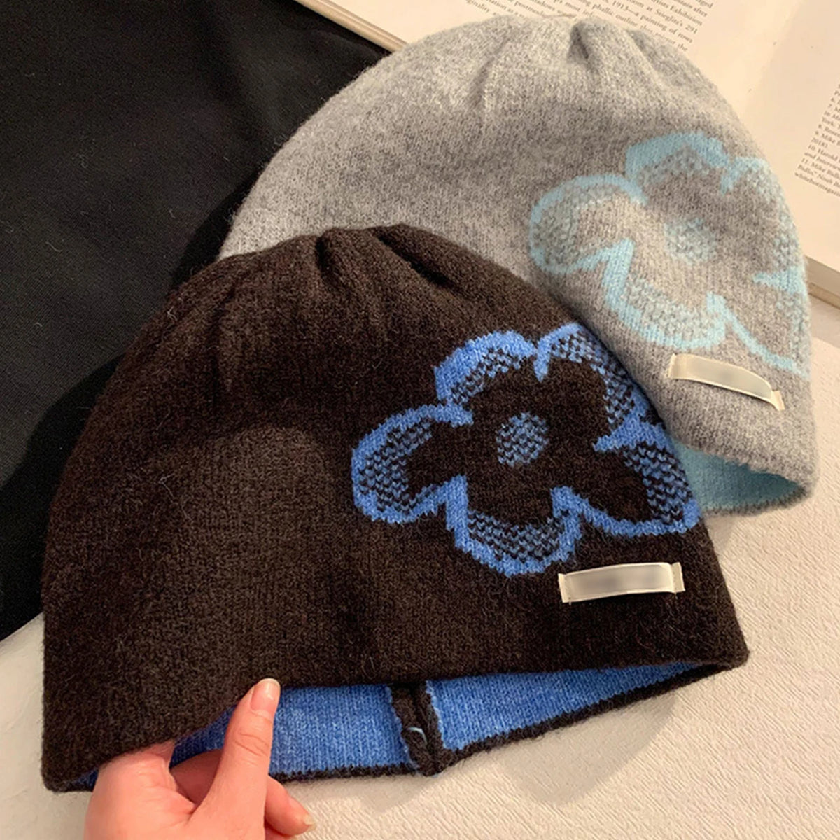 Three knit beanie hats are shown in a styled product shot: one dark brown, one light gray, and one black, each with a large blue flower motif and a small white tag. A hand is holding the front brown beanie.