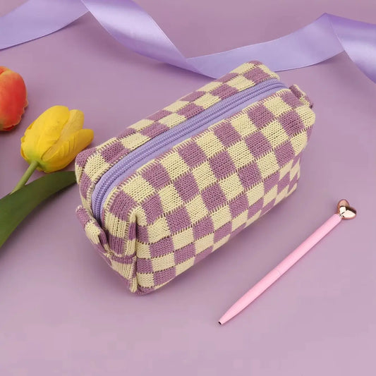 A small zippered fabric pouch with a lavender and cream checkerboard pattern, styled on a lavender background with a pink pen, tulips, and a purple ribbon.