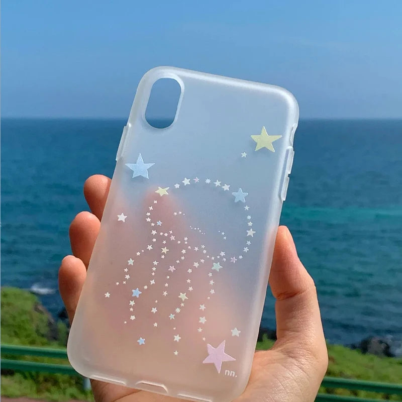 A hand holds a translucent frosted phone case decorated with pastel star and jellyfish designs, set against a blurred background of the blue ocean and sky.