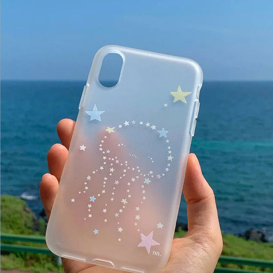 A hand holds a translucent frosted phone case decorated with pastel star and jellyfish designs, set against a blurred background of the blue ocean and sky.