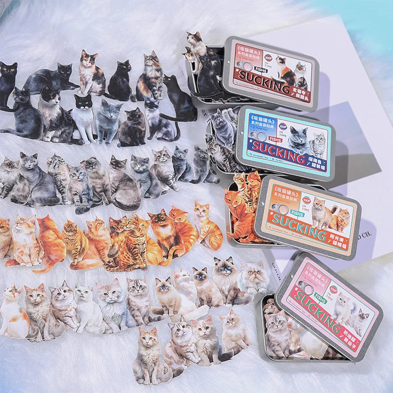 A styled flat-lay of cat-themed stickers and matching metal tins on a white faux-fur surface. The stickers feature various breeds of cats in different poses and colors, including black, white, gray, orange, and tabby.
