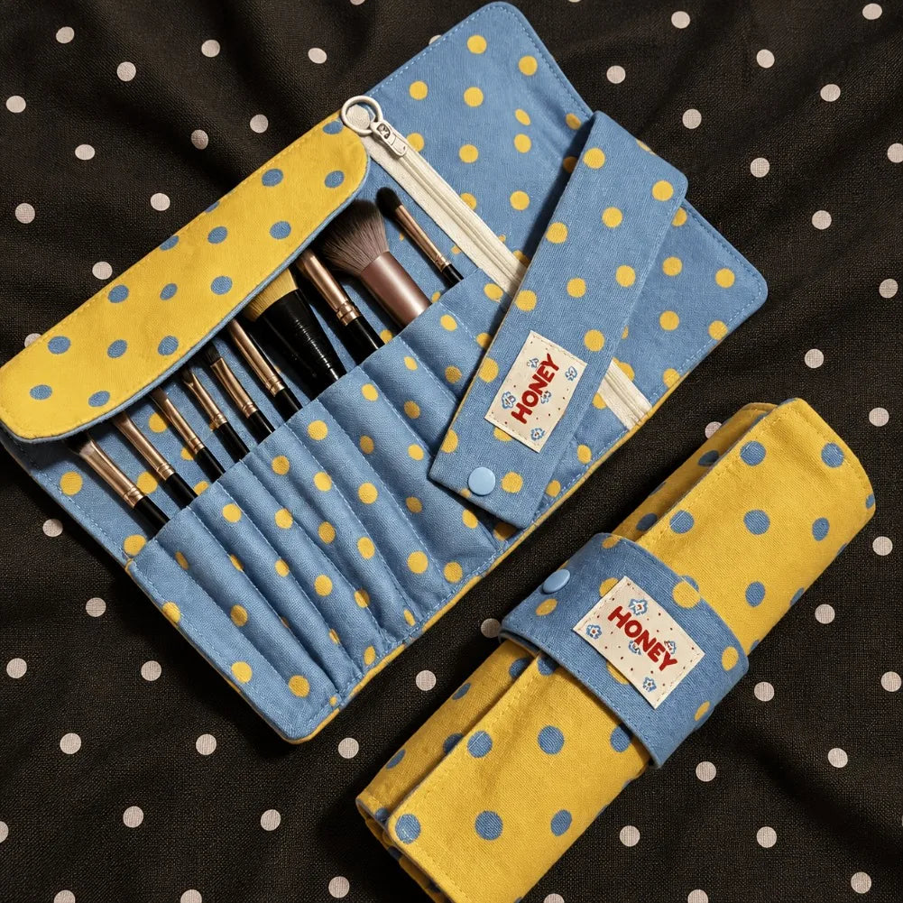 A blue and yellow polka dot makeup brush roll organizer shown open with several makeup brushes inside, plus a matching rolled-up version placed below it on a black fabric background with white polka dots. A fabric tag on both pieces reads HONEY.
