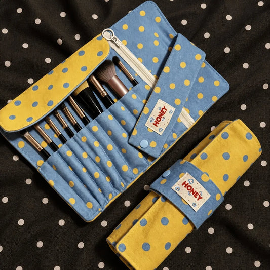 A blue and yellow polka dot makeup brush roll organizer shown open with several makeup brushes inside, plus a matching rolled-up version placed below it on a black fabric background with white polka dots. A fabric tag on both pieces reads HONEY.