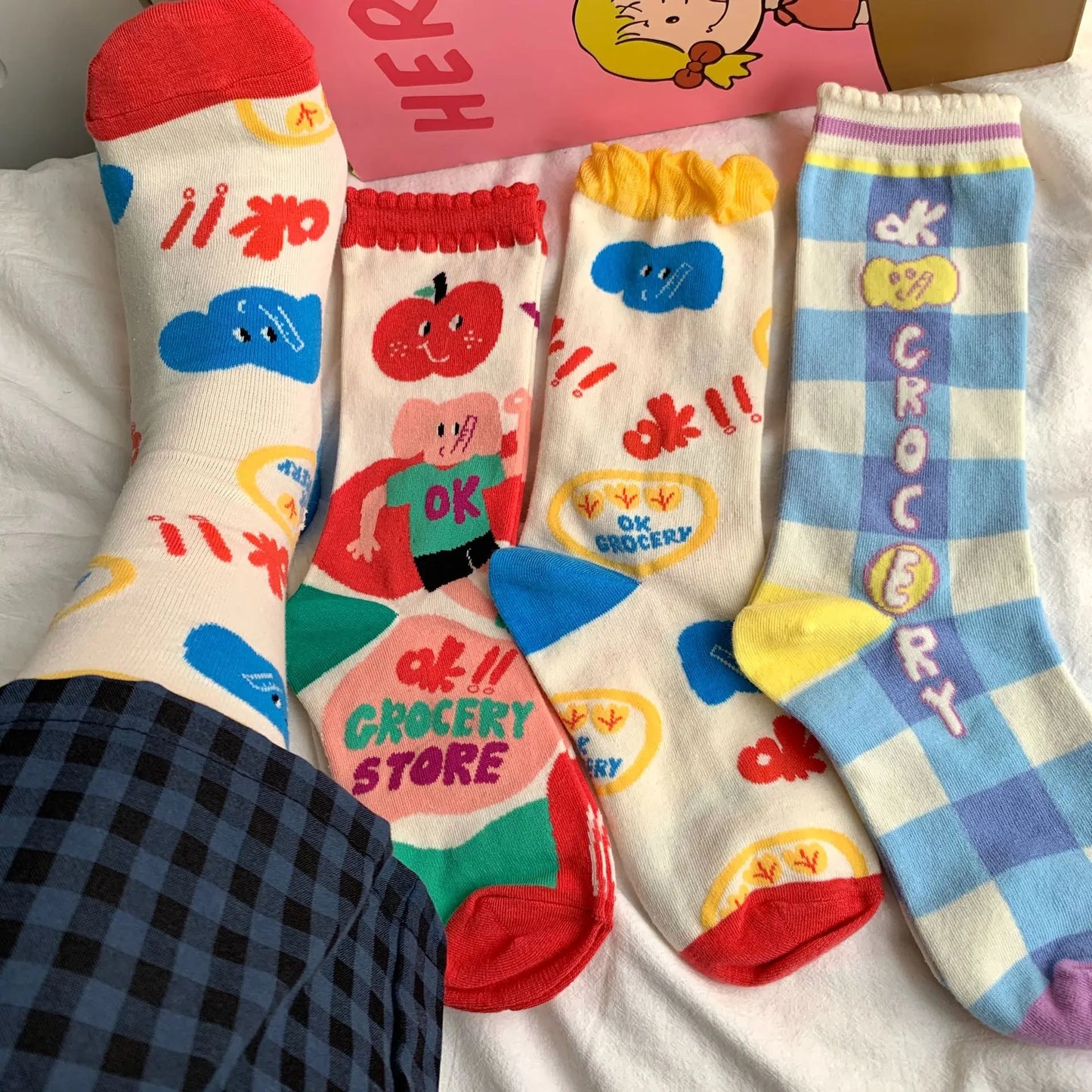 Four novelty crew socks laid flat on a white fabric surface, each with a different colorful cartoon grocery-themed print including clouds, fruit, a character, and blue-and-white stripes with vertical text. A pink box and part of a plaid garment are visible in the background and foreground.