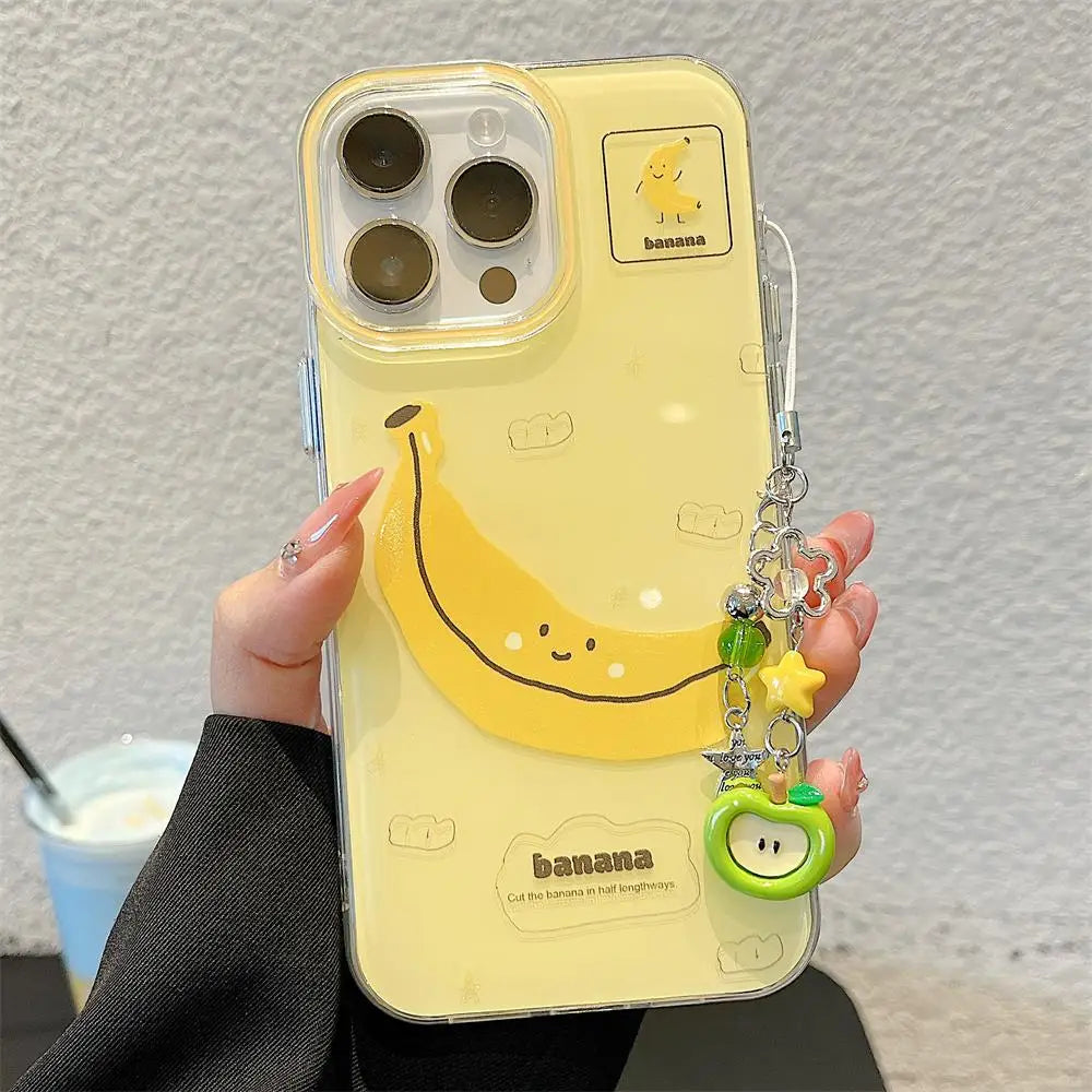 A hand holds a pale yellow phone case with a large smiling banana graphic, a small banana icon near the camera cutout, and a dangling charm with beads and a green apple-shaped accent. The case appears to be shown on a phone, photographed against a light textured wall with a drink partially visible in the background.