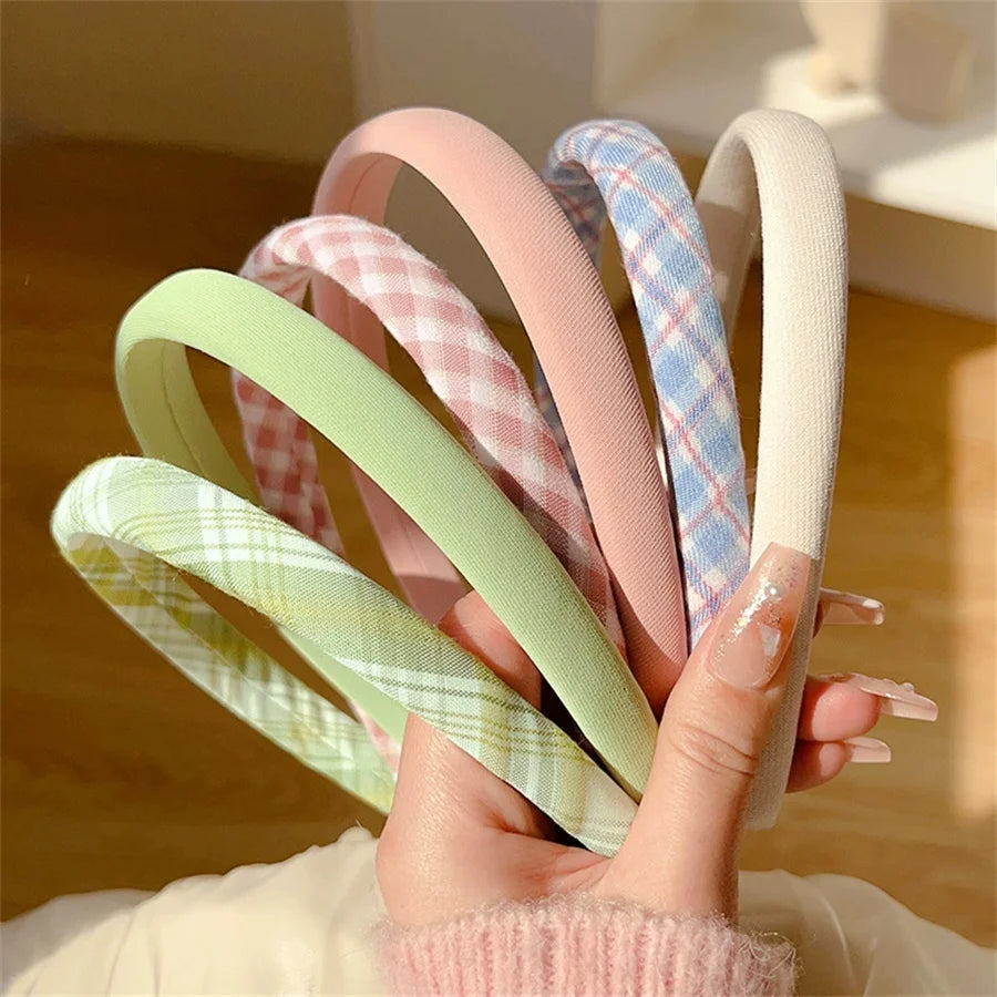 A hand holds a fan of six wide fabric headbands in pastel colors, including pink, light green, blue plaid, and cream. The headbands have a soft woven texture, and the background shows a warm, softly lit indoor setting.