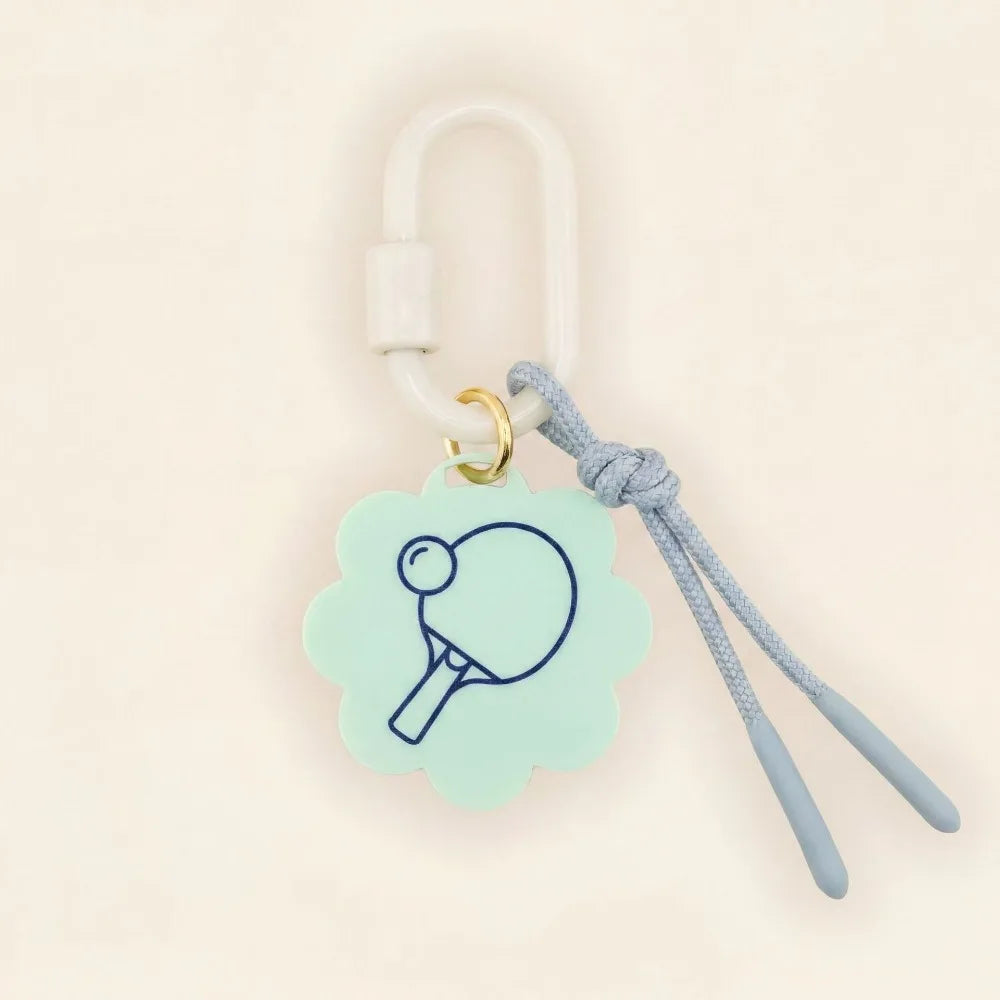 A light mint green flower-shaped charm featuring a navy blue ping-pong paddle graphic, attached to a white carabiner, a light blue cord, and a small gold ring, set against a warm cream background.