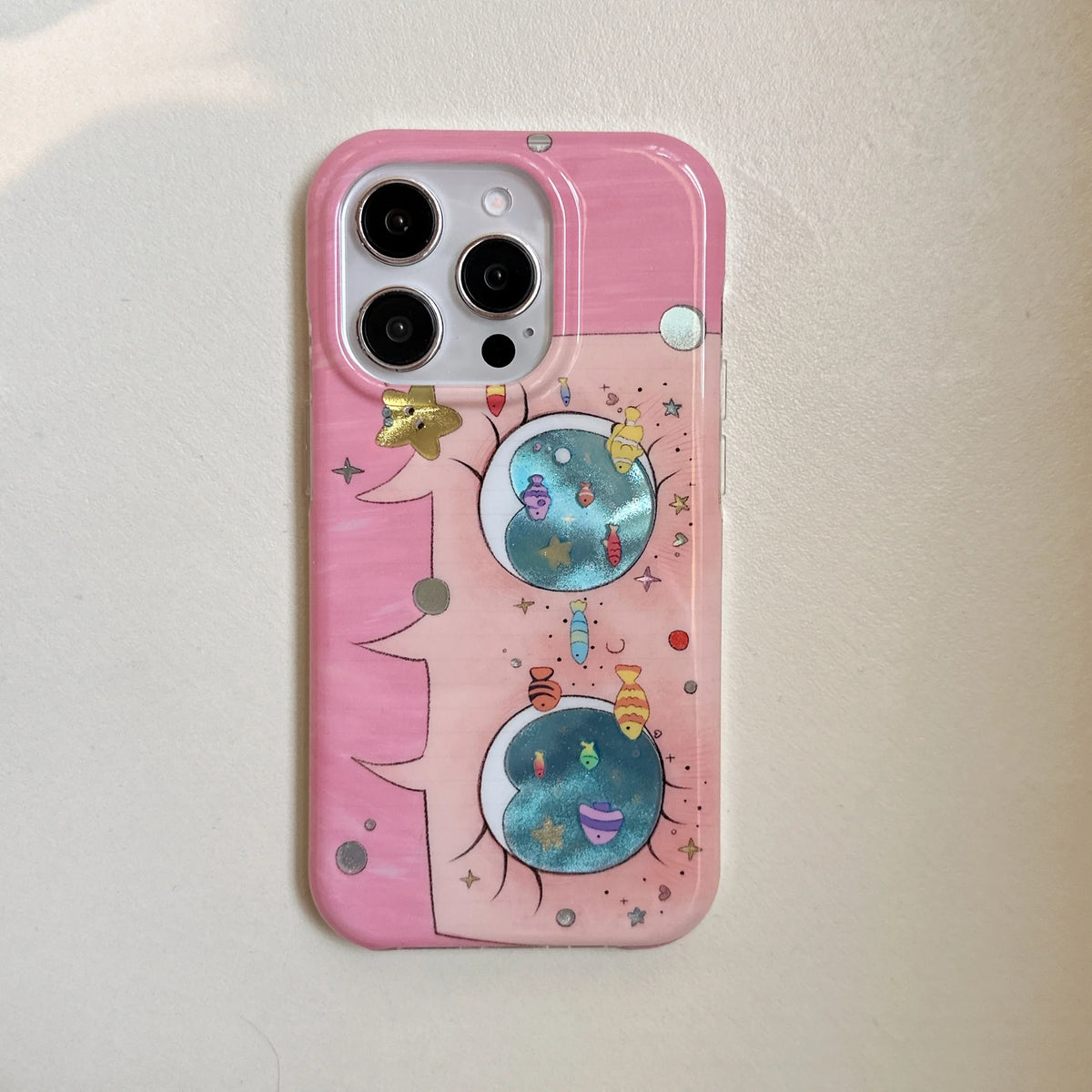 A pink phone case with a whimsical cartoon design featuring two smiling blue planets, stars, and small colorful celestial elements, shown on a light beige textured background.