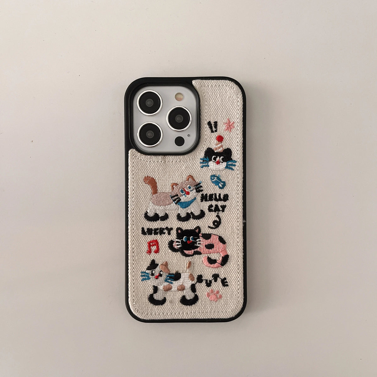 A beige canvas phone case with black trim and colorful embroidered cat illustrations and text, shown on a light gray background.