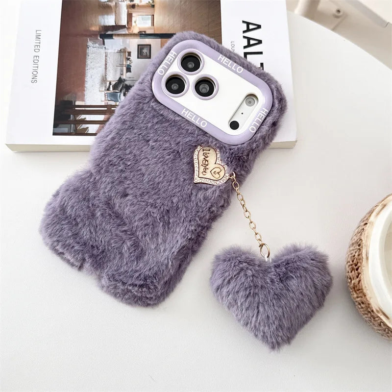 A plush lavender purple phone case with a matching fuzzy heart charm and gold chain, displayed on a white surface next to a book.