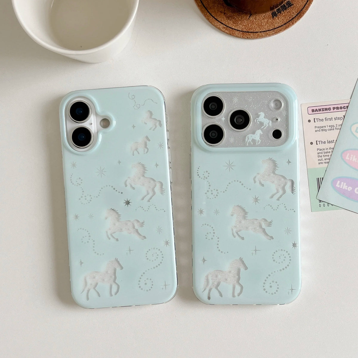 Two light blue phone cases with embossed unicorn designs, displayed on a white surface with a white mug, a cork coaster, and a sticker partially visible.