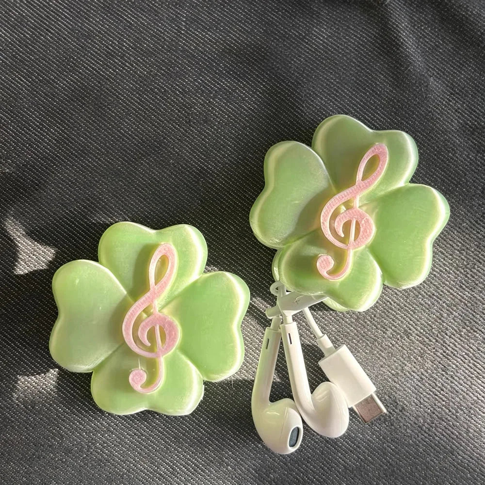 Two light green four-leaf clover-shaped accessories with pink treble clef designs, shown with white wired earphones on a dark textured fabric background.