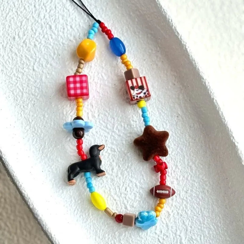 A colorful beaded phone strap or necklace featuring various themed wooden beads, including a dachshund, star, football, gingham, and popcorn pieces, displayed on a textured white surface.