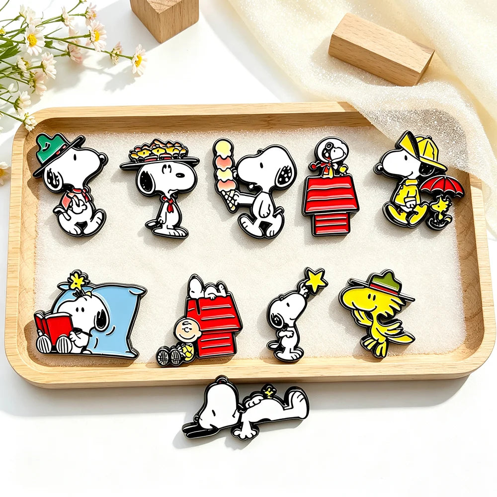 A set of Snoopy and Woodstock enamel pins arranged on a light wood tray with a few scattered pins below, photographed on a clean white surface with soft fabric and small flowers as styling props.