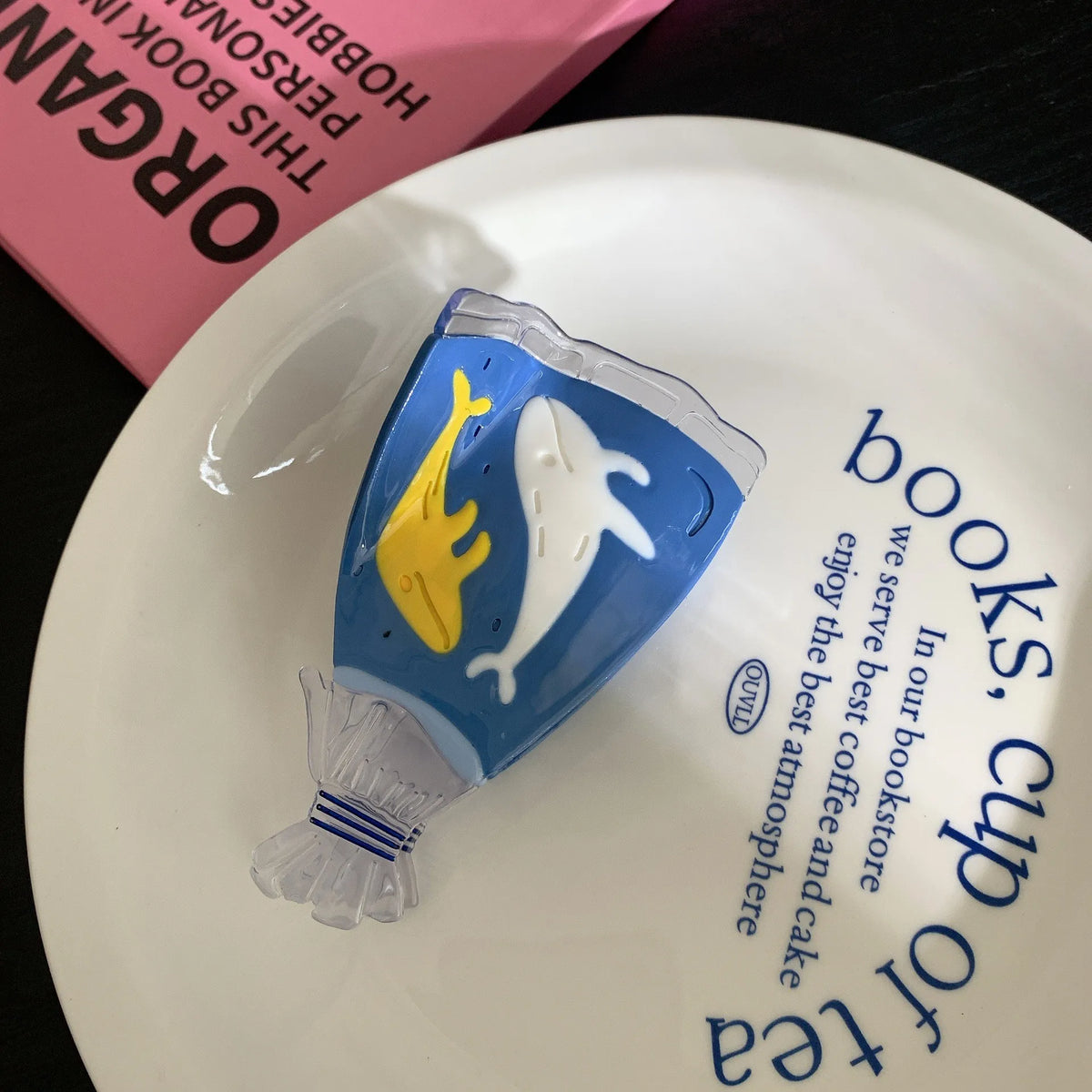 A novelty cup-shaped object with a blue body, white dolphin, and yellow fish design, wrapped in clear plastic and resting on a white plate with blue text.