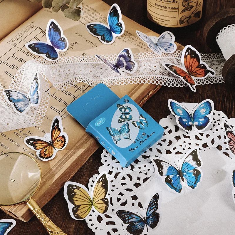 A blue box of butterfly-shaped stickers or decals lies on a white lace doily over an open vintage book, surrounded by scattered butterfly cutouts in blue, orange, purple, and yellow on a wooden surface.