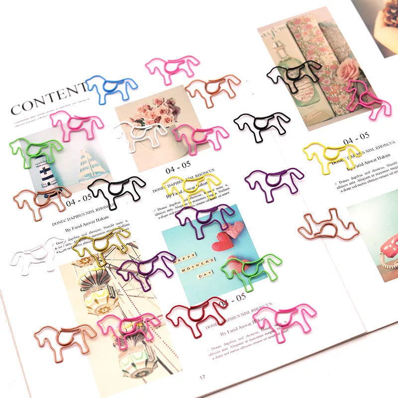 A set of colorful horse-shaped metal paperclips arranged on an open magazine page. The clips come in multiple colors including pink, green, blue, yellow, purple, black, and rose gold, with some showing a small heart detail on the tail.