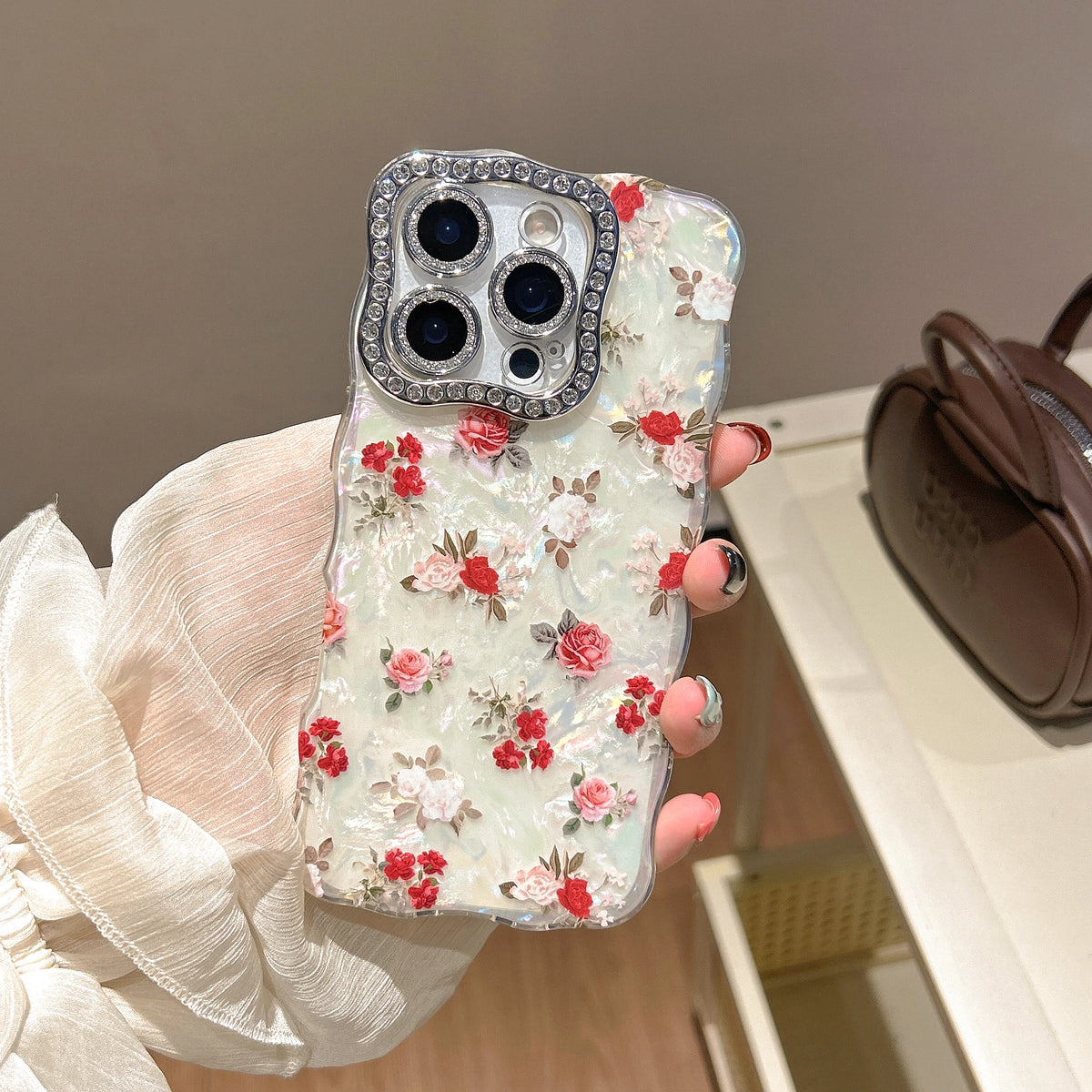 A hand wearing a white sheer sleeve holds a clear phone case decorated with red and pink 3D roses on a light green background, featuring a rhinestone-embellished camera frame.
