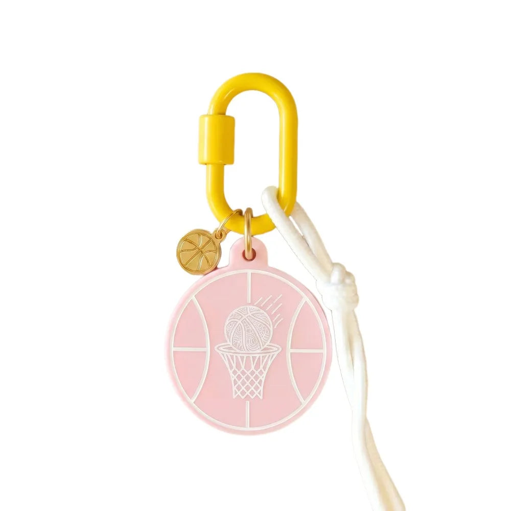 A pink circular basketball-themed keychain or bag charm with a white rope and a yellow carabiner, shown on a plain white background.