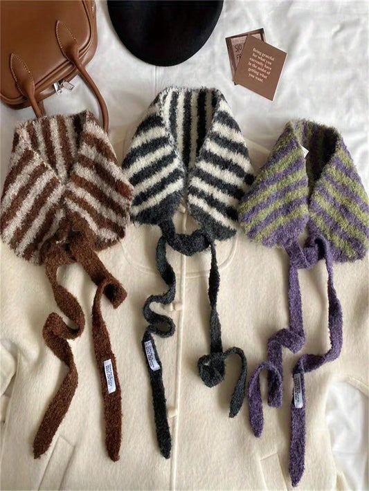 Three fuzzy knit tie-neck scarves or shawls laid flat on a cream fabric surface: one brown and cream striped, one black and cream striped, and one purple and olive striped. A brown handbag corner, a black hat, and two small cards are partially visible around them.