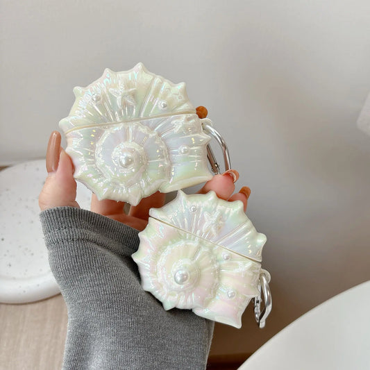 A hand wearing a gray long-sleeve shirt holds two iridescent white earbud cases decorated with 3D floral and pearl designs, set against a soft, neutral indoor background.