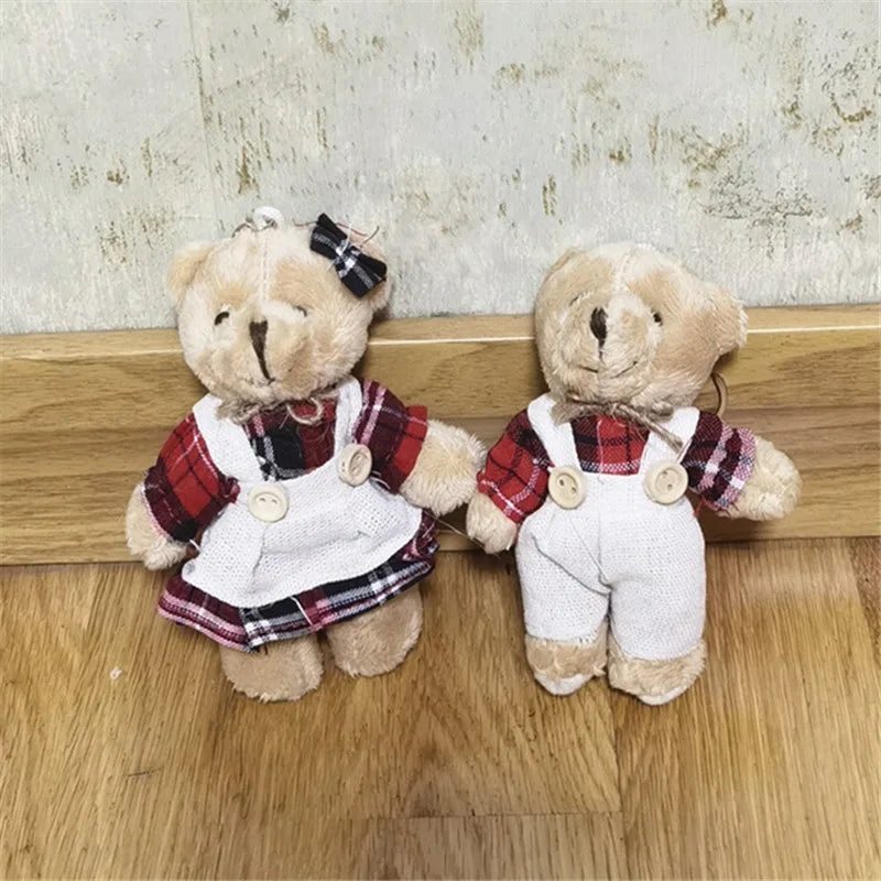 Two small teddy bear plush dolls sitting side by side on a wooden surface against a textured wall. One wears a red plaid shirt with a white pinafore dress and a black bow, and the other wears a matching red plaid shirt with white overalls.