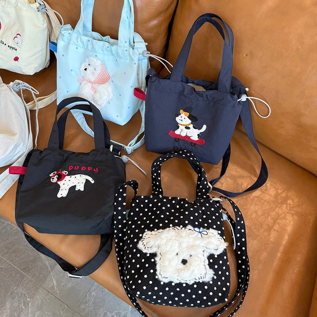 Four small dog-themed tote bags arranged on a brown leather couch. The bags include white, light blue, navy, and black fabrics with white polka dots and embroidered or applique dog designs.