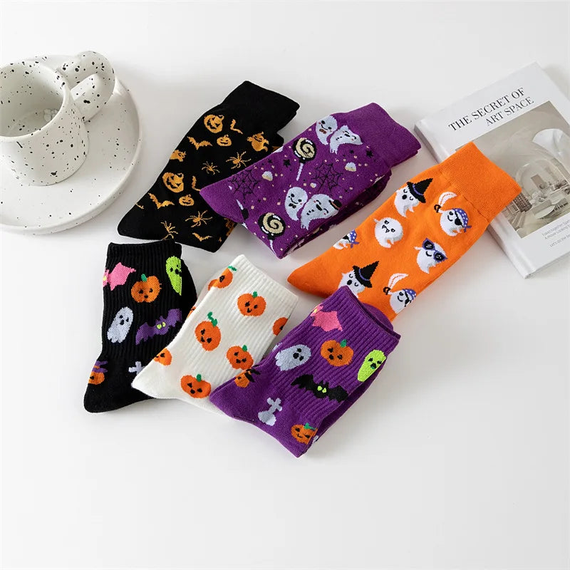 Six pairs of Halloween-themed socks in black, purple, orange, and white, featuring cute ghost, pumpkin, witch, and bat patterns, arranged on a white surface with a teacup and a book.