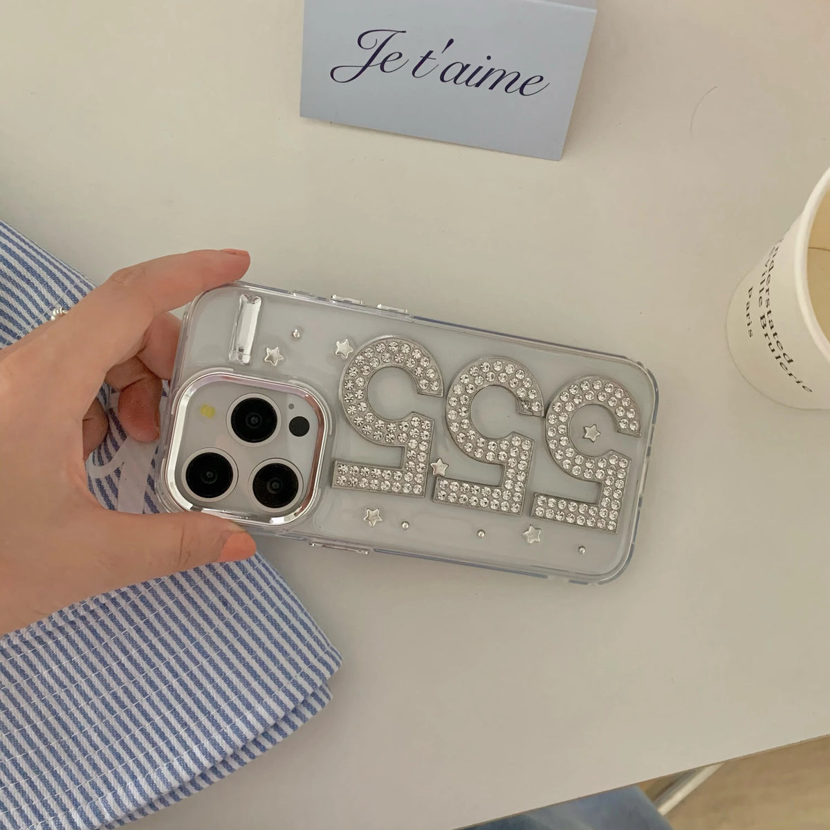 A hand holds a clear phone case decorated with large rhinestone-embellished '555' numbers and small star details, shown on a light tabletop with a blue striped cloth and a small card in the background.