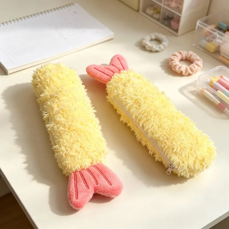 Two fuzzy yellow pencil cases shaped like shrimp, with pink tails, displayed on a white desk alongside stationery accessories.