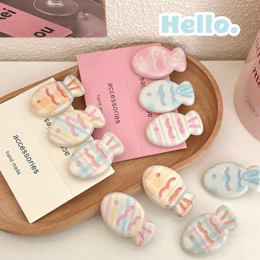 A set of pastel fish-shaped hair clips with striped patterns in pink, blue, yellow, and orange, displayed on a wooden tray with pink and cream cards labeled 'handmade accessories'.