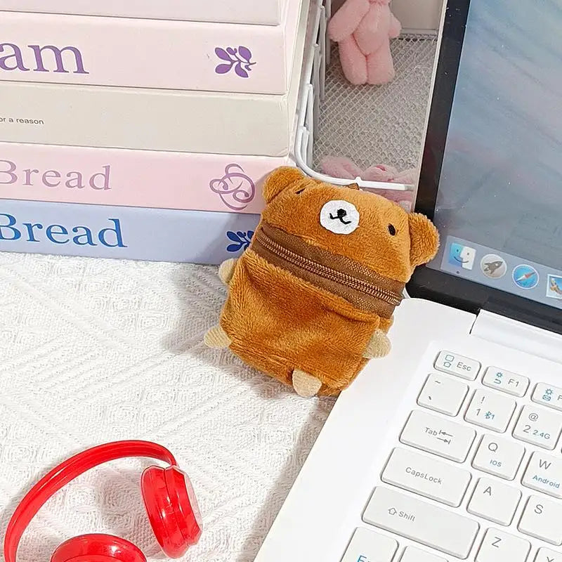 A small plush bear-shaped zippered pouch in tan and brown, placed on a white textured surface next to a white laptop keyboard, with pink books and a red pair of headphones in the background.