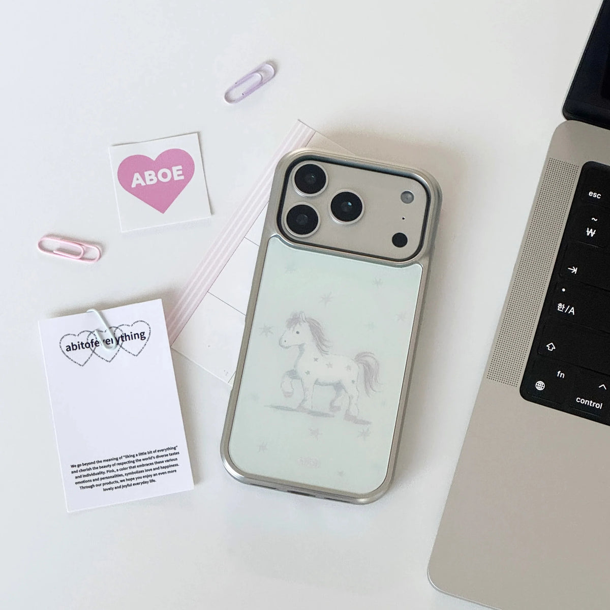 A phone case with a silver metallic frame and a pale mint back featuring a whimsical horse illustration, styled on a white desk next to a laptop keyboard with paper clips and cards.