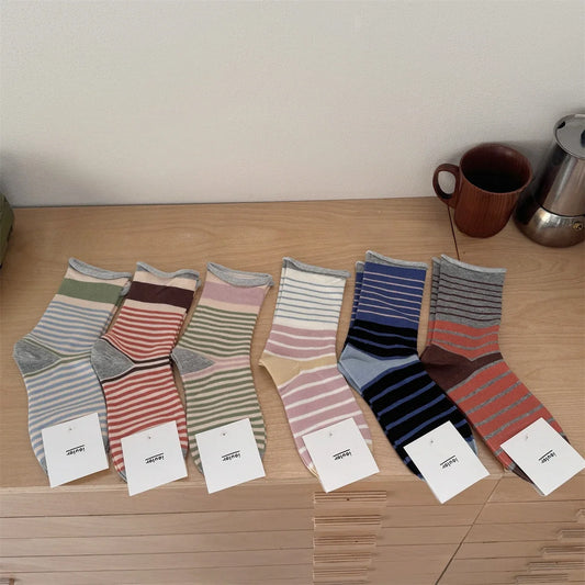 Six pairs of striped socks arranged on a light wood surface against a plain wall, each with a small white tag attached. The socks come in muted stripe combinations including blue-gray, pink-beige, olive green, rust red, navy, and gray-brown.