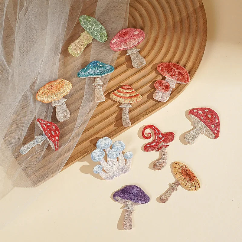 A set of embroidered mushroom patches in various colors and patterns, arranged on a beige surface with a wooden tray and sheer tulle fabric.