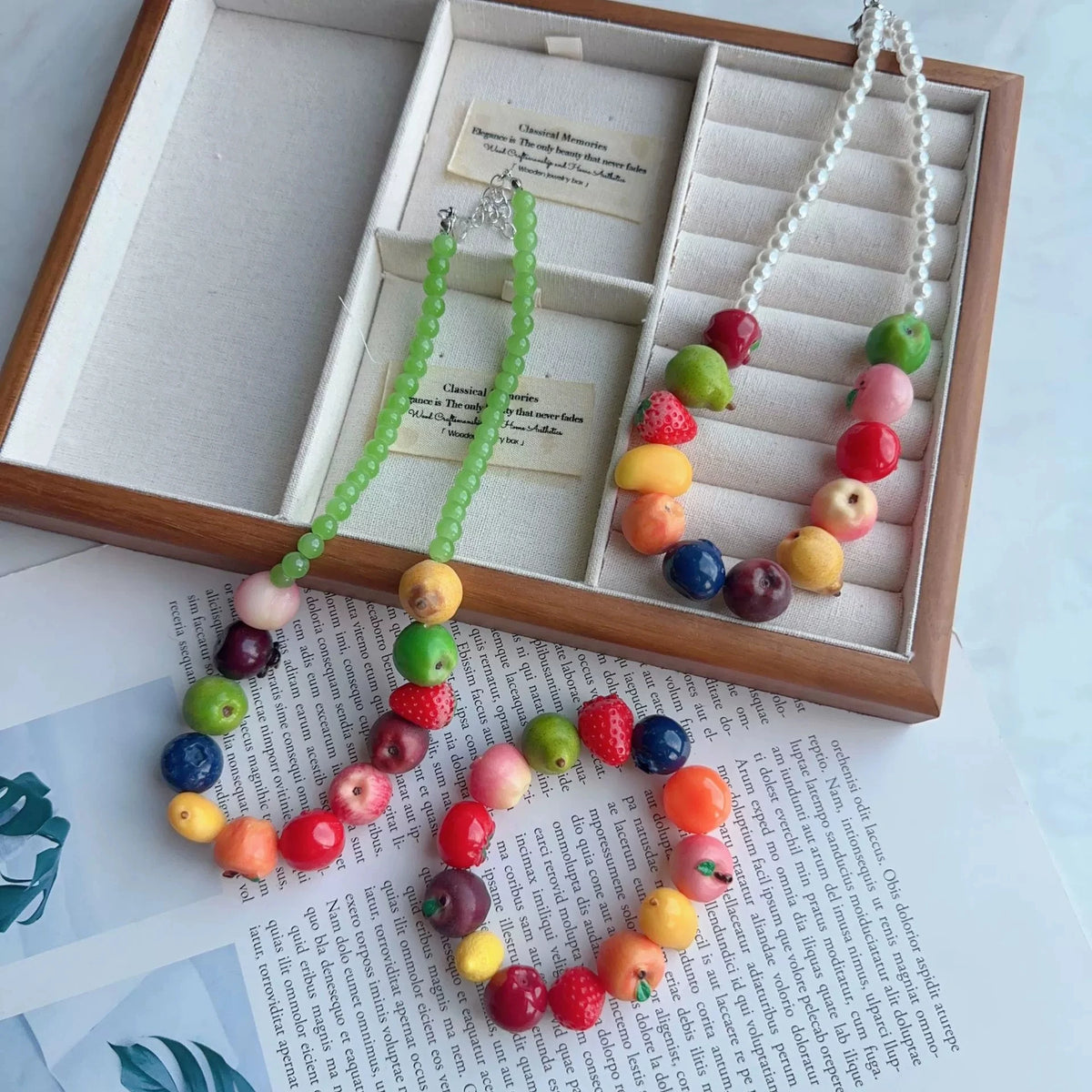 Three colorful beaded necklaces and matching bracelets displayed inside a wooden jewelry box, with printed papers underneath on a light surface.