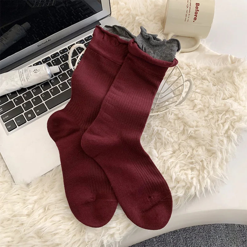 A pair of ribbed crew socks in burgundy, with one sock folded over to show a gray ruffled inner lining, styled on a white faux fur surface next to a laptop, a mug, and a tube of cream lotion.