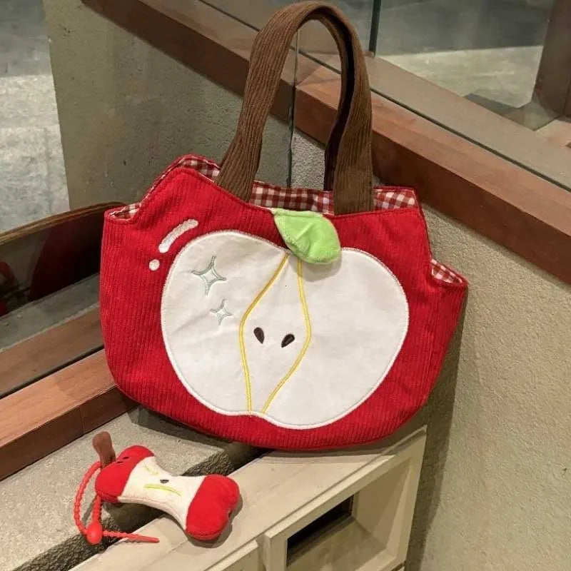 A red corduroy tote bag shaped like a stylized apple, with brown handles, a white apple face motif, and a small green leaf accent, displayed indoors on a shelf next to a matching small plush accessory.