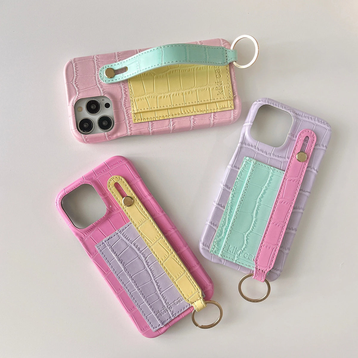 Three crocodile-embossed phone cases arranged on a light gray background, each with a pastel color-blocked back panel and a wrist strap keychain. The top case is pink and mint, the bottom left is pink and lavender with yellow, and the right case is lavender and mint.