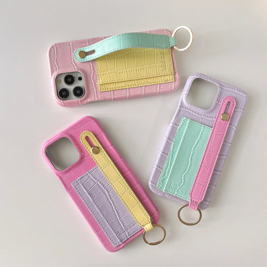 Three crocodile-embossed phone cases arranged on a light gray background, each with a pastel color-blocked back panel and a wrist strap keychain. The top case is pink and mint, the bottom left is pink and lavender with yellow, and the right case is lavender and mint.