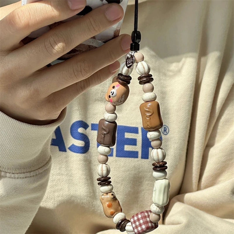 A close-up of a phone strap with cute cookie and pastry charms, worn around the neck over a cream sweatshirt with blue lettering.