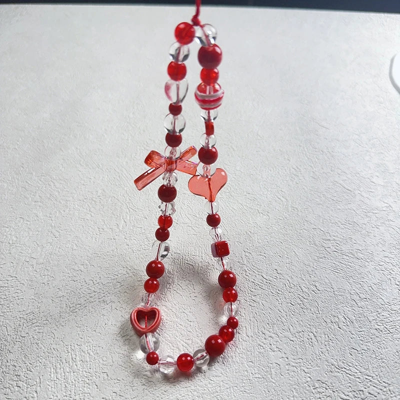 A red and clear acrylic beaded phone strap with heart and bow charms, displayed on a textured light gray surface.