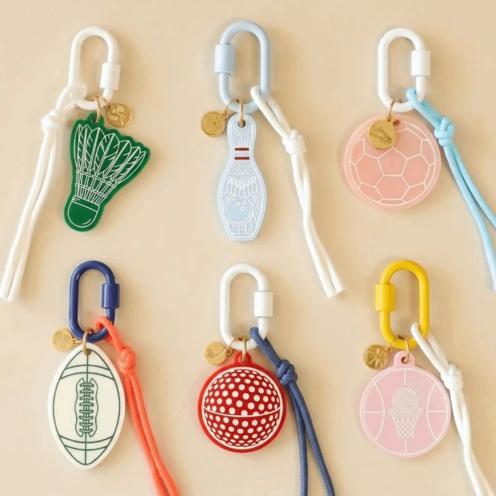 Six colorful silicone sports keychains with white carabiners and cords, arranged in a grid on a beige background. The designs include a green badminton shuttlecock, a light blue bowling pin, a pink soccer ball, a blue football, a red basketball, and a pink basketball with an ice cream cone graphic.